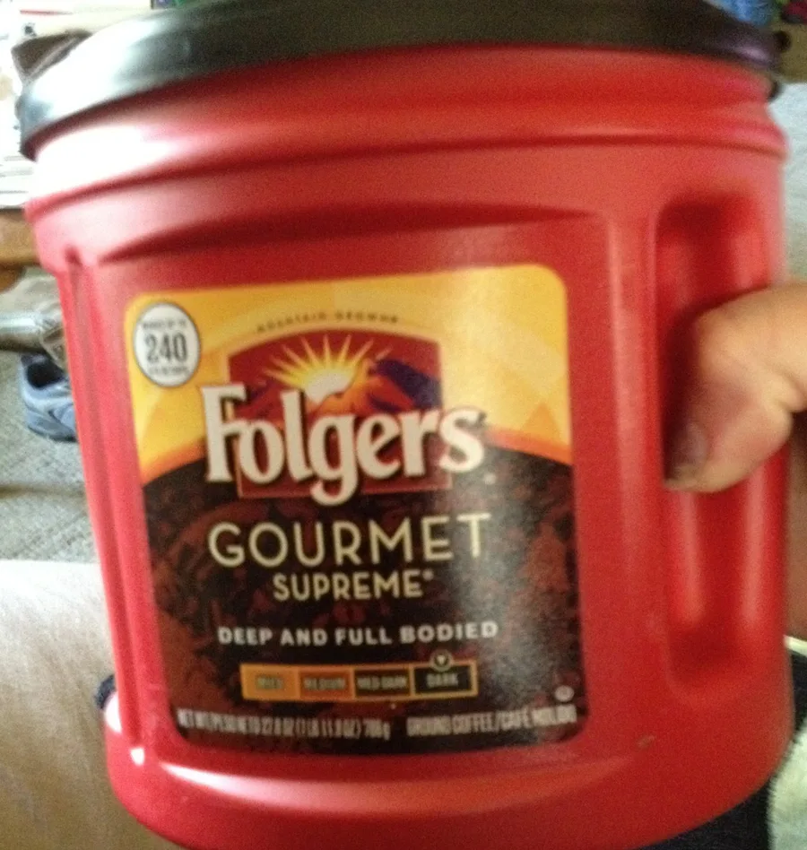 A hand holding a red Folgers Gourmet Supreme coffee can