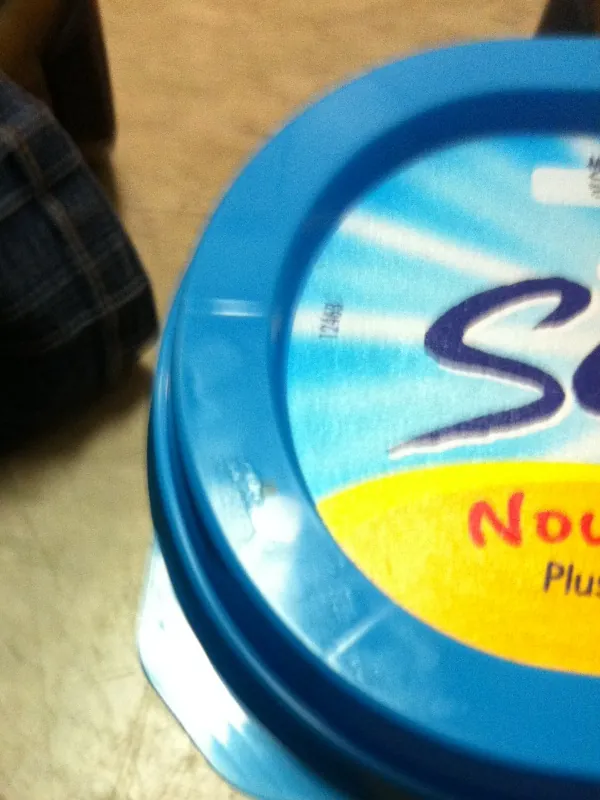 The blue lid of a yogurt tub on a floor next to a pair of jeans; no date is printed on the visible part